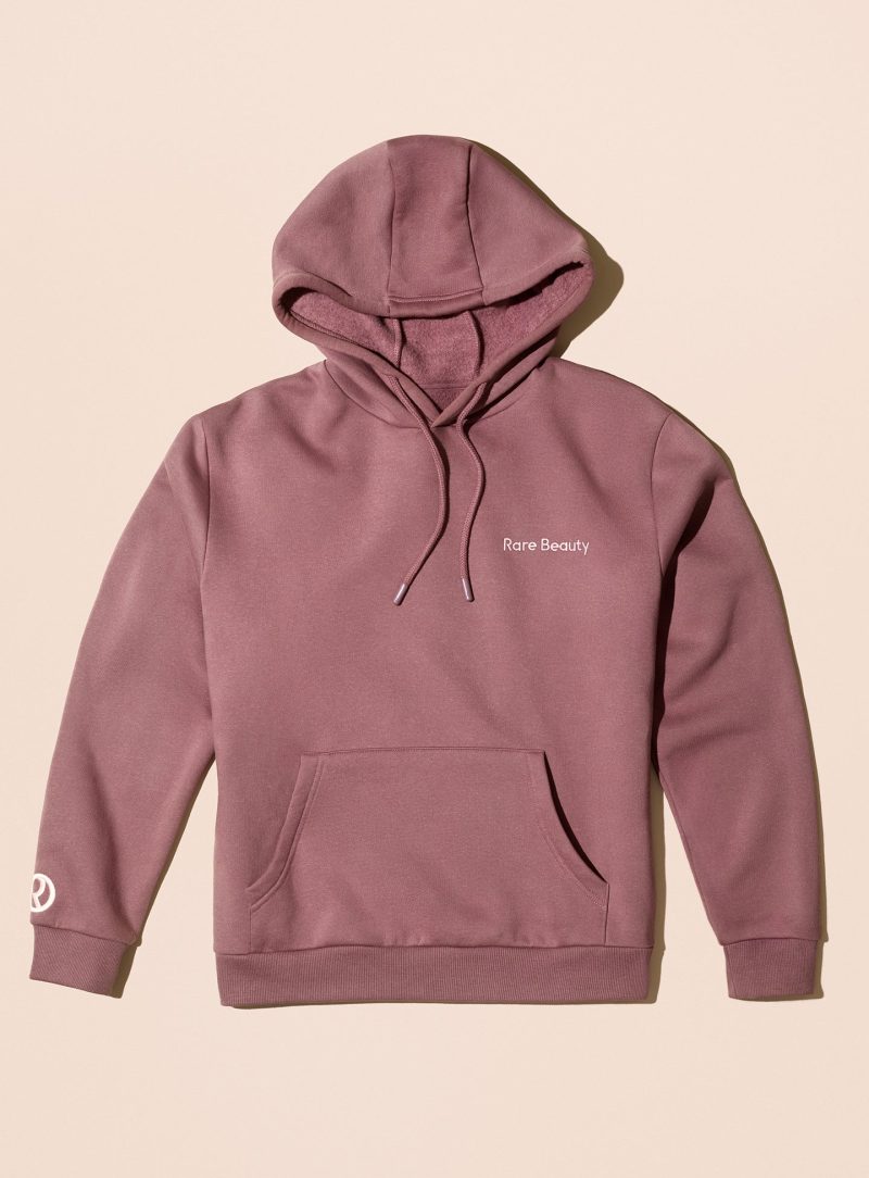 Rare Beauty's Comfy Hoodie mauve hooded sweatshirt laying against a neutral beige background.